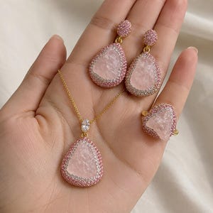 May include: A jewelry set consisting of a necklace, a pair of drop earrings, and a ring. Each piece features a central pale pink stone with a textured surface, surrounded by a border of small pink crystals set in a gold-toned metal frame.