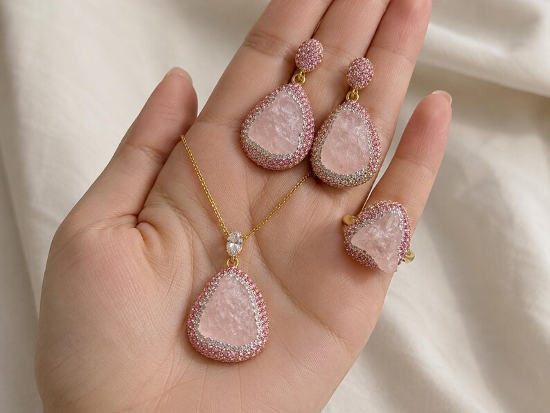 May include: A jewelry set consisting of a necklace, a pair of drop earrings, and a ring. Each piece features a central pale pink stone with a textured surface, surrounded by a border of small pink crystals set in a gold-toned metal frame.