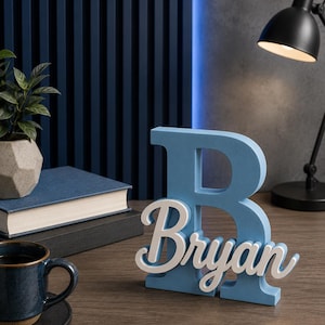 May include: A light blue wooden letter "B" with the name "Bryan" in white script. The personalized decor sits on a wooden desk, with a dark blue mug, books, and a small plant in the background. A black desk lamp is also visible.