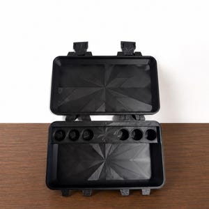 May include: A black rectangular storage case with a hinged lid and two locking latches. The interior features a geometric faceted pattern on the base and lid, with a horizontal insert containing six circular compartments for small items.