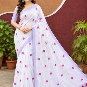 May include: A white saree featuring small pink floral embroidery throughout the fabric. The garment is finished with a light purple border and matching purple tassels along the bottom edge. The fabric has a textured appearance and light drape.