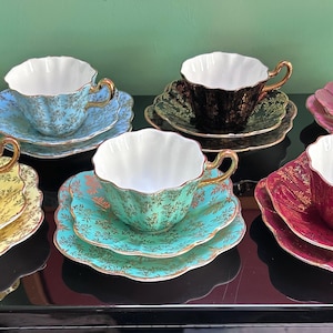 May include: Six vintage teacups and saucers in a range of colours: yellow, blue, green, pink, and dark red. Each teacup has a scalloped edge and gold trim, sitting on matching saucers. The teacups are displayed on a reflective surface.