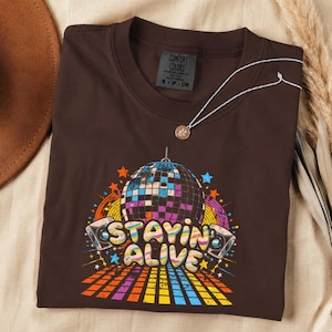 May include: A dark brown short-sleeve t-shirt featuring a graphic print of a disco ball, two speakers, and a colorful grid floor. The text "STAYIN ALIVE" is printed in a wavy, multi-colored font. The shirt has a Comfort Colors brand neck label.