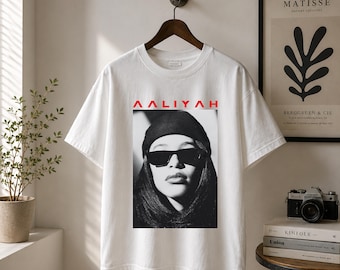 Peut inclure: Un t-shirt blanc à manches courtes avec une impression photographique en noir et blanc représentant une personne portant un bonnet et des lunettes de soleil. Le nom AALIYAH est imprimé en lettres capitales rouges au-dessus de l'image.