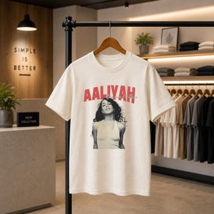 May include: A cream-colored short-sleeve t-shirt featuring a black and white photographic print of Aaliyah. The text AALIYAH is printed in large red capital letters above the image, with the script text One in a Million below it.