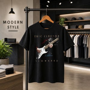 May include: A black short-sleeve t-shirt featuring a graphic print of a person playing a white electric guitar. The text "ERIC CLAPTON" appears above the guitar, and the word "SLOWHAND" is printed below the graphic in a light-colored font.