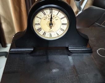Könnte beinhalten: Eine schwarze Kaminuhr mit gewölbtem Oberteil und rechteckigem Sockel. Das Zifferblatt ist cremefarben mit schwarzen römischen Ziffern, schwarzen Zeigern und dem Text "VICTORIA STATION 1747 LONDON" in der Mitte mit goldenen Verzierungen.