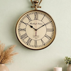 May include: A round wall clock with a brass-colored frame and a pocket watch-style top handle. The off-white face features black Roman numerals, black hour and minute hands, a red second hand, and the text Huntleigh Station Cambridge England 1924.