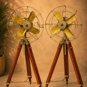 May include: Two floor fans featuring four gold-toned metal blades and a circular wire cage. Each fan is mounted on a dark brown wooden tripod stand with brass hardware and a small chain connecting the legs. The centre of each fan displays a circular label reading Made for Royal Navy London.