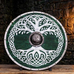 May include: A circular wooden shield featuring a dark green finish and a white painted tree of life design with intricate knotwork patterns. The shield has a metallic rim secured with rivets and a central domed metal boss in the middle.
