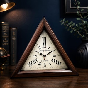 Peut inclure: Une horloge triangulaire en bois avec un cadran crème, des chiffres romains noirs et des aiguilles noires avec une trotteuse rouge. Le cadran porte les inscriptions Jones & Hendricks EST 1923 et OXFORD. Le cadre est en bois brun foncé.