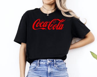 May include: Black t-shirt with the Coca-Cola logo in red. The classic script logo is centered on the front of the shirt. The shirt is paired with blue denim shorts.