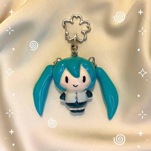 May include: A keychain featuring a small, glossy, chibi-style figure with bright teal hair styled in long twin tails. The figure has a white and dark teal outfit and is attached to a silver-colored metal clasp shaped like a five-petaled cherry blossom.