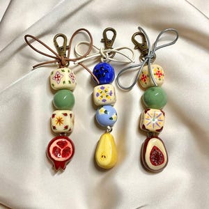 May include: Three decorative keychains with colourful beads and fruit-shaped charms. Each keychain features a different design, including floral patterns, a pomegranate, a pear, and a fig. The keychains are attached to gold-coloured clasps.