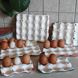 Peut inclure: Un ensemble de cinq plateaux à œufs en céramique blanche de tailles variées, chacun doté d'un bord festonné et de petits motifs de cœurs rouges ou verts. Les plateaux sont disposés pour contenir des œufs de poule bruns et blancs.