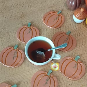 Könnte beinhalten: Eine weiße Keramikteetasse gefüllt mit dunklem Tee und einem Silberlöffel, umgeben von sieben terrakottafarbenen kürbisförmigen Untersetzern mit grünen Stielen und braunen Linien sowie zwei kleinen dekorativen Kürbisfiguren.