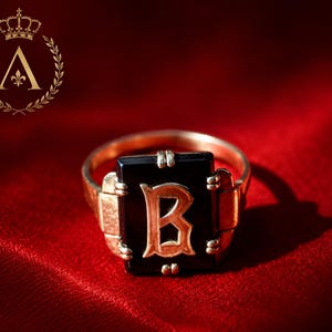 May include: A gold ring featuring a rectangular black onyx stone set in the centre. The stone is inlaid with a prominent gold letter B in a gothic-style font. The ring band is made of polished gold and has a simple, clean design.
