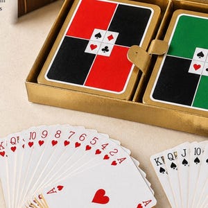 May include: A vintage playing card set in a gold box. The cards feature red, black, and green designs. The box includes the text "Waddingtons" and "Card Games". A fanned display of playing cards with red hearts is in the foreground.