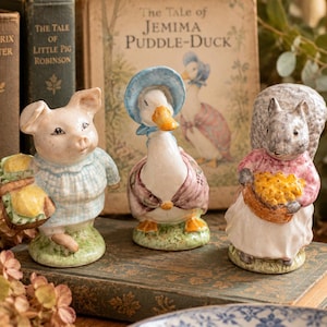 May include: Three ceramic figurines featuring a pig in a blue checkered outfit holding a basket, a duck wearing a blue bonnet and shawl, and a squirrel in a pink top and white skirt holding a basket of yellow items, all on green bases.