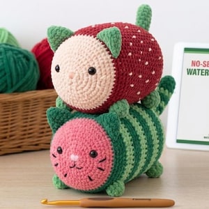 May include: Two crocheted cat-shaped plushies, one resembling a strawberry and one a watermelon, stacked vertically. A tablet displays the text NO-SEW CAT LOAF PATTERNS: WATERMELON & STRAWBERRY next to a basket of yarn and a single crochet hook.