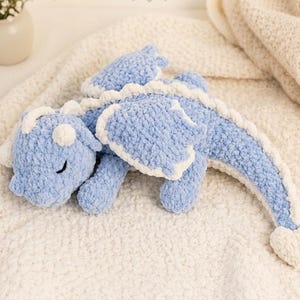 May include: A light blue crocheted dragon plush toy with white accents on the wings, spine, and head. The dragon is lying on a cream-colored blanket. The dragon has a smiling face and is a soft toy.