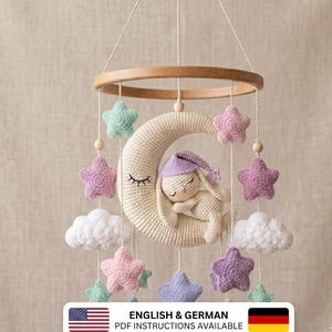 Puede incluir: Un móvil de ganchillo con una luna creciente de color crema con cara dormida, un pequeño conejo crema con gorro de dormir morado sobre la luna, y estrellas de peluche colgantes en menta, rosa y morado, además de nubes blancas y cuentas de madera.