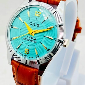 May include: A wristwatch with a turquoise dial, gold-tone hands, and gold-tone numerals 12 and 6. The dial features the text ORIS, 17 JEWELS, SHOCK PROOF, and SWISS MADE. It has a silver-tone fluted bezel and a textured brown leather strap.