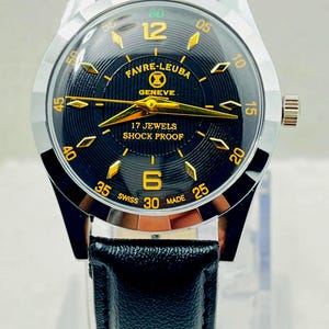 Può includere: Un orologio da polso con quadrante nero testurizzato, lancette color oro e indici color oro. Il quadrante riporta la scritta FAVRE-LEUBA GENEVE, 17 JEWELS, SHOCK PROOF e SWISS MADE. Presenta una cassa in metallo color argento e un cinturino in pelle nera.