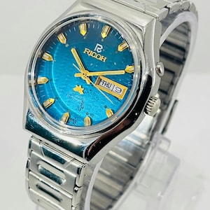 Op de afbeelding: Een Ricoh-polshorloge met een metallic blauwe wijzerplaat, goudkleurige wijzers en goudkleurige uurmarkeringen. Het horloge heeft een dag-datumvenster op de drie-uurpositie, een gouden sterembleem en een zilverkleurige roestvrijstalen schakelarmband.