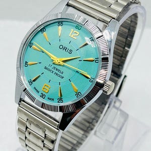 Peut inclure: Une montre-bracelet argentée avec un cadran turquoise à texture côtelée, des aiguilles dorées et des chiffres dorés à 6 et 12 heures. Le cadran affiche le texte ORIS, 17 JEWELS, SHOCK PROOF et SWISS MADE. Elle possède une lunette cannelée et un bracelet en métal.