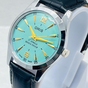 Op de afbeelding: Een polshorloge met een turquoise wijzerplaat, goudkleurige wijzers en goudkleurige uurmarkeringen. De wijzerplaat bevat de tekst ORIS, 17 JEWELS, SHOCK PROOF en SWISS MADE. Het heeft een zilverkleurige metalen kast en een zwarte lederen horlogeband.