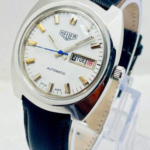 Op de afbeelding: Een vintage Heuer automatisch polshorloge met een zilverkleurige roestvrijstalen kast en een witte wijzerplaat. Het horloge heeft goudkleurige uurmarkeringen, een blauwe secondewijzer en een dag-datumvenster met 2 TUE 18. Zwarte lederen band.