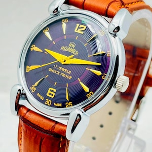 Può includere: Un orologio da polso Roamer con cassa rotonda color argento e quadrante viola testurizzato. Il quadrante presenta lancette e indici color oro, con la scritta RW ROAMER, 17 JEWELS, SHOCK PROOF e SWISS MADE. Dotato di cinturino in pelle marrone.