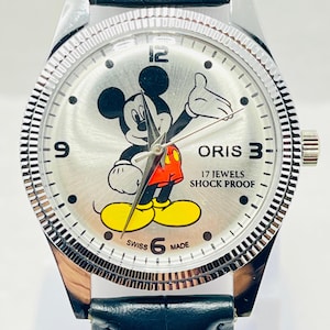 May include: A silver-toned analogue wristwatch featuring a Mickey Mouse graphic on the dial, black numerals at 3, 6, 9, and 12, and the text ORIS, 17 JEWELS, SHOCK PROOF, and SWISS MADE. The watch has a fluted bezel and a black leather strap.