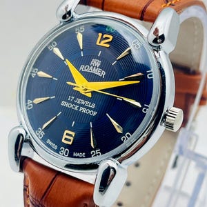 Può includere: Un orologio da polso Roamer con cassa rotonda color argento, quadrante blu scuro con lancette e indici dorati, e la scritta 17 JEWELS SHOCK PROOF e SWISS MADE. L'orologio include un cinturino in pelle marrone con motivo a coccodrillo testurizzato.