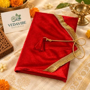 Puede incluir: Una cubierta rectangular de terciopelo rojo con flecos metálicos dorados en los bordes y un cordón dorado con una borla. Una tarjeta blanca con el texto VEDAVIBE NATURALS y un logotipo de hoja verde se encuentra junto al objeto.