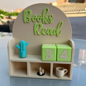 Puede incluir: Un estante de exhibición beige en forma de arco con el texto "Books Read" en letras verdes en relieve. Incluye dos cubos numéricos verdes que muestran "34", una figura de cactus azul brillante, tres velas blancas pequeñas y una taza beige.