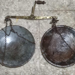 Könnte beinhalten: Eine antike Waage mit zwei runden Metallschalen, die an Ketten hängen. Die Waage hat einen Messingbalken und einen Holzgriff. Die Schalen und Ketten weisen Alters- und Abnutzungserscheinungen auf.