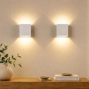 May include: Two square white wall sconces mounted on a beige wall, emitting warm light upwards and downwards. Below, a light-coloured vase with green leaves sits on a wooden surface next to a small stack of books and two small ceramic bowls.