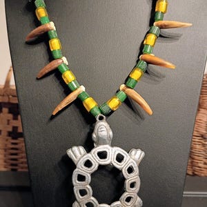 May include: A necklace featuring a silver-toned metal turtle pendant with a hollow centre. The strand consists of alternating green and yellow cylindrical beads, interspersed with black and white patterned beads and several curved brown spikes.