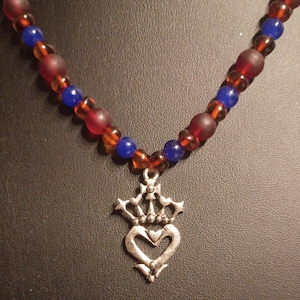 May include: A necklace featuring a silver-toned metal pendant shaped like a crown atop a heart. The strand consists of alternating round beads in translucent amber, matte dark red, and smooth blue, creating a colorful and textured pattern.