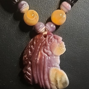 May include: A necklace featuring a carved purple and cream shell pendant in a profile silhouette. The pendant is strung on a cord with alternating round yellow beads, small purple striped beads, and larger black beads with white etched patterns.