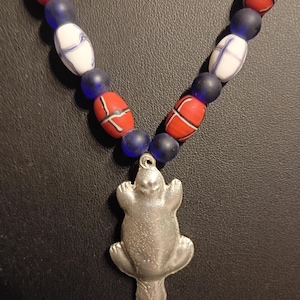 May include: A necklace featuring a silver-toned metal pendant shaped like a platypus. The strand consists of alternating matte blue spherical beads and oval-shaped beads in red and white, each decorated with thin black cross-hatch line patterns.