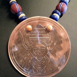 May include: A circular copper pendant featuring an engraved spider design with a central cross motif and two small spherical accents at the top. The pendant is suspended from a necklace strand of alternating red, blue, and white spherical beads.