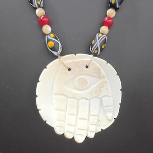 May include: A necklace featuring a large, circular, off-white carved pendant depicting a hand with an eye in the center. The cord is strung with black beads containing yellow and white patterns, interspersed with small, round, translucent red and beige beads.