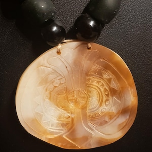 May include: A necklace featuring a large, round, light brown carved shell pendant with intricate geometric patterns and a central cross design. The pendant is suspended from a strand of smooth, matte black spherical beads of varying sizes.