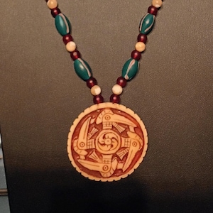 May include: A necklace featuring a round, tan-colored pendant with a carved, dark-red, bird-like motif. The chain consists of alternating dark-red, cream, and teal-colored beads, with the teal beads displaying a subtle, light-colored linear pattern.