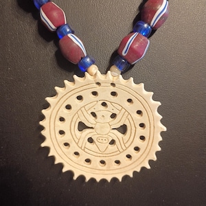 May include: A necklace featuring a circular, off-white pendant with a serrated edge and a central spider engraving, surrounded by a ring of small holes. The strand consists of alternating dark red beads with white stripes and translucent blue beads.