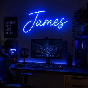 Pode incluir: Um letreiro de néon azul brilhante exibindo o nome James em uma fonte cursiva, montado em uma parede acima de uma mesa de computador. O letreiro emite um brilho vibrante, iluminando o ambiente da sala de jogos com luz azul fria.