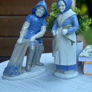 Może przedstawiać: Dwie porcelanowe figurki przedstawiające rybaka i sprzedawczynię ryb. Rybak ma na sobie niebieski kapelusz, niebieską górę i szare buty, trzymając sieć. Sprzedawczyni ryb ma niebieskie nakrycie głowy, niebieską górę i biały fartuch, trzymając rybę.